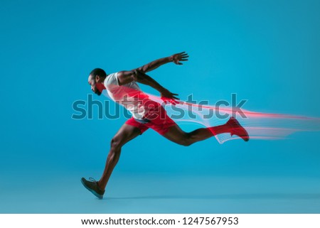 Full length portrait of active young african muscular running man, isolated over blue studio background with flashes of light