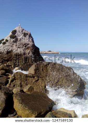 Spalashes of water on sidebank's rocks. And a gull seating on top of one rock. Clear sky and rumbling tide.  Royalty-Free Stock Photo #1247589130