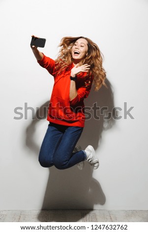 Full length photo of lovely woman 20s wearing red sweatshirt jumping and taking selfie on cell phone isolated over white background
