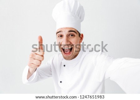 Happy chef cook wearing uniform standing isolated over white background, taking a selfie, giving thumbs up