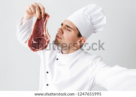 Cheerful chef cook wearing uniform showing raw beef steak isolated over white background, taking a selfie