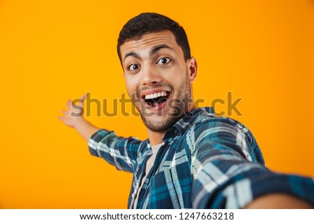 Cheerful young man wearing plaid shirt standing isolated over orange background, taking a selfie