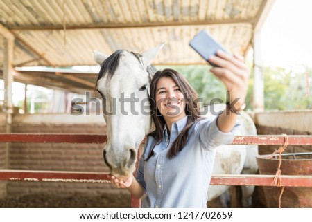 Happy good looking woman using smartphone to make selfie with white horse at stable