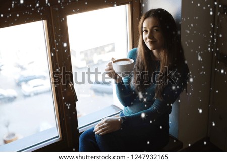 Cute girl sitting with a cup of hot cocoa or coffee by the window in winter with snow background.