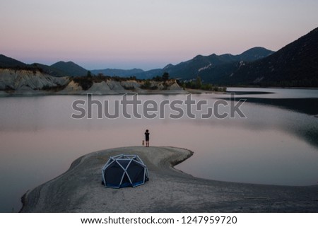 Man stands next to camping tent on lake or seaside beach, makes inspirational photo on smartphone of sunset light. Best friend, trail adventure dog or puppy next to him enjoy outdoors