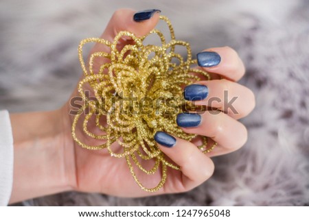Girl with navy blue manicure on finger nails holding decorative golden flower on gray background. Manicure concept image. Close up, selective focus