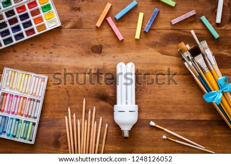 Creative art idea fantasy imagination concept, light bulb, paint brushes, paintbox with watercolors, crayons and pencils on brown wooden background, artistic table desk. Top view, flat lay copy space