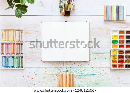 Paint brushes set, paintbox with watercolors, crayons, pencils and open notebook paper on white wooden background, artistic creative art workplace accessories. Top view, flat lay with copy space