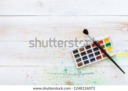 Learning painting concept, paint brush and box with watercolors on white wooden table with splashes, artistic background, creative art workplace for children kids, top view with copy space, flat lay