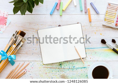 Creative workplace concept, flat style artistic design table, blank notebook mock up for writing drawing sketches, pencil, paintbrushes set and cup of coffee on white wooden desk, top view copy space