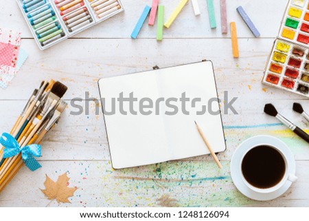Flat style artistic design workspace, blank notebook mock up for creative sketches with watercolor paints, pencil, set of paintbrushes and cup of coffee on white wooden table, top view, copy space