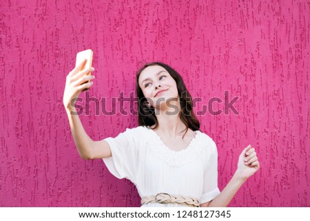 Girl taking selfie on modern smartphone. Lovely young woman looking at display of cellphone with gladness and showing victory sign by hand. Isolated on pink