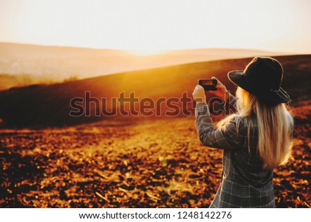 Back view of anonymous woman in stylish outfit using smartphone to take pictures of magnificent countryside during sunset.Unrecognizable female taking photos of evening countryside