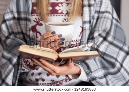 Girl drinking hot tea and reading book in long winter night at home. Woman warming and covered with retro blanket. Cold weather and winter concept. Close up, selective focus
