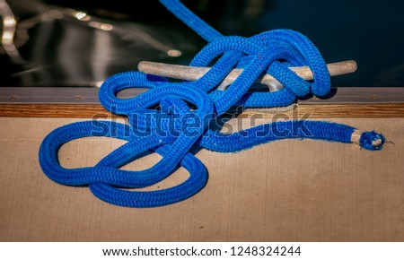 A blue nylon mooring rope tied around a cleat, with visible water in the background.  The rope has flecks of red and white in it.
