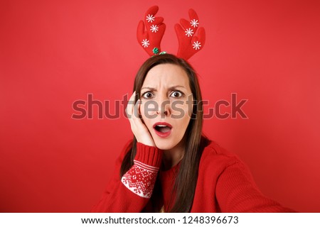 Close up selfie shot of puzzled young Santa girl in fun decorative deer horns put hand on cheek isolated on red background. Happy New Year 2019 celebration holiday party concept. Mock up copy space