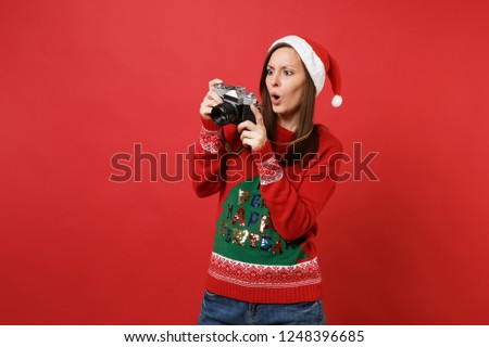 Shocked young Santa girl taking pictures on retro vintage photo camera, keeping mouth wide open isolated on red background. Happy New Year 2019 celebration holiday party concept. Mock up copy space