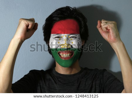 Cheerful portrait of a man with the flag of Tajikistan painted on his face on grey background. The concept of sport or nationalism. Red white and green with a crown surmounted by stars.