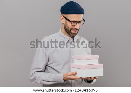 Indoor shot of handsome unshaven man with brislte holds two boxes, happy to recieve present from friend on birthday, wears festive clothes, models against grey background. Young male with gift