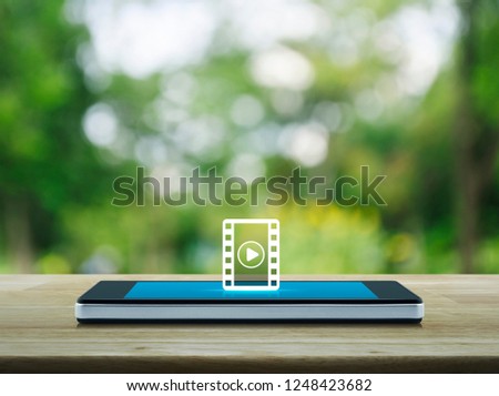 Play button with movie flat icon on modern smart mobile phone screen on wooden table over blur green tree in park, Business cinema online concept