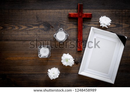 Funeral. Mockup of portrait of the deceased, of dead person. Frame with black ribbon near flowers, candles and cross on dark wooden background top view copy space