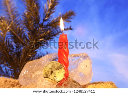 New Year's decoration. Bitcoin sunk into the ice and brightened by the flame of a red candle against the background of Christmas tree branches and a blue sky. The freezing of financial assets.