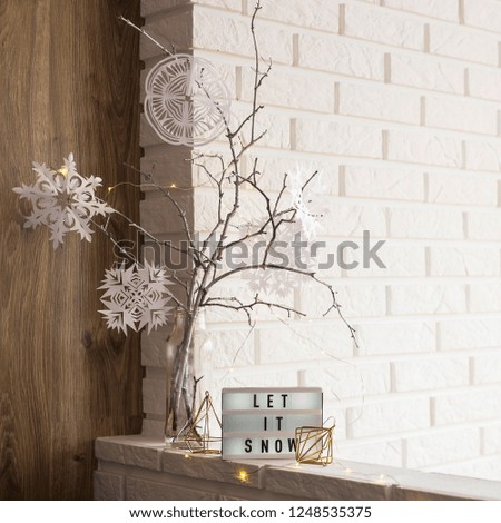 Let it snow it is written on a decorative lamp next to a home winter decor with a vase with tree branches with paper snowflakes against a brick wall of an apartment