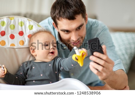 family, fatherhood and people concept - father with little baby daughter in highchair taking selfie by smartphone at home