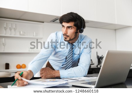 Image of a concentrated young business man at the kitchen wearing headphones work with documents using laptop computer.