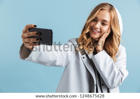 Cheerful young girl wearing raincoat standing isolated over blue background, taking a selfie
