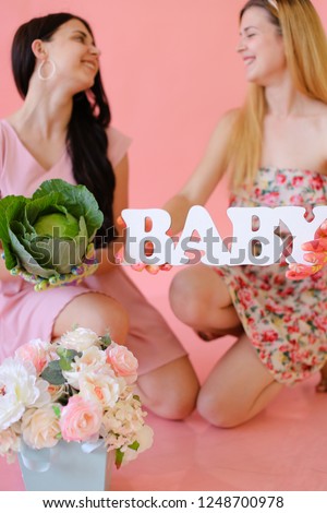 Young female persons keeping cabbage and inscription baby in pink monophonic backgrpund. Concept of photo session for pregnant mom.