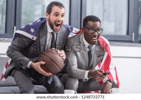 happy multicultural business partners with american flag watching basketball match in office