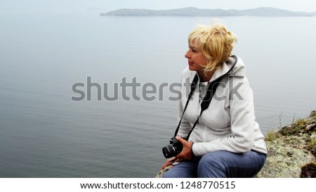 A woman in sports clothes sits on the stone of a rocky cliff. The traveler with photo camera enjoys a walk along the sea coast. The concept of a healthy and active lifestyle in senior adults.
