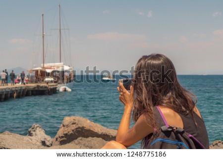 A young woman taking a picture in her vacations on the islands of the Mediterranean sea. A wedding is undergoing on the pier of the island. 