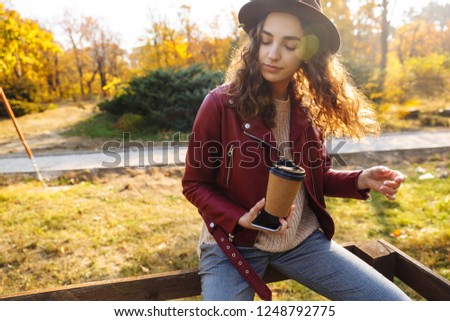 Picture of a young curly woman sitting in the autumn park holding coffee and mobile phone.