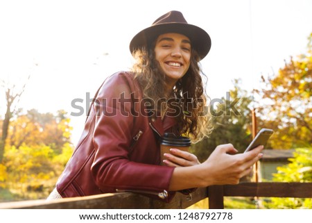 Picture of a young curly woman walking in the autumn park drinking coffee using mobile phone.