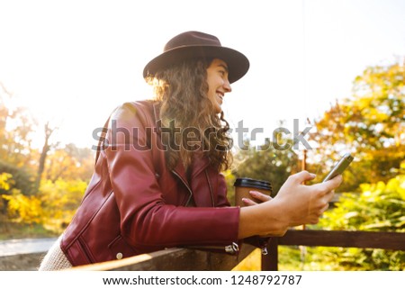 Picture of a laughing curly woman walking in the autumn park drinking coffee using mobile phone.