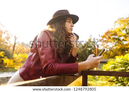 Picture of a young curly woman walking in the autumn park drinking coffee using mobile phone.
