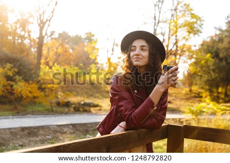 Picture of a beautiful young cute curly woman walking in the autumn park drinking coffee.