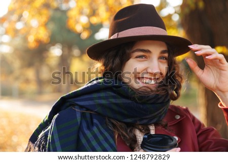 Picture of a beautiful young cute curly woman walking in the autumn park drinking coffee.