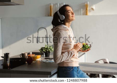 Image of an amazing happy healthy woman in the kitchen standing daily morning routine eat salad listening music in headphones.