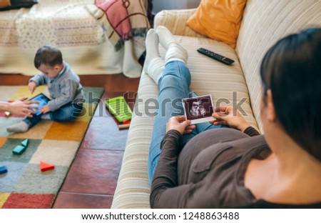 Pregnant woman looking at the ultrasound of her new baby while the older brother plays