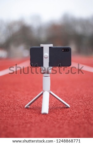 Mock up of a black smartphone with a tripod. White screen for design template on the background of the red track for running and sports stadium