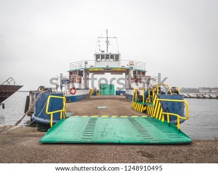 Perspective view of the small littoral ferry, which is moored with open ramp and ready for boarding passengers and cars. Symmetrical panorama with cloudy sky and empty ship with antennas on flybridge.