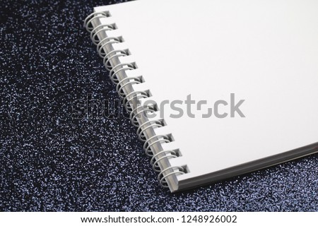 White spiral notebook with clean sheets on black glittery lighted background, side view.