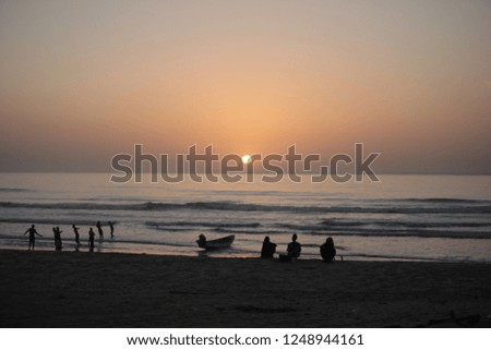  a group of african boys pulling a fishing net out of a water on Atlantic beach during bright sunset, with a big boat on a shore, and a group of people sitting on the sand, outdoors