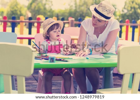 young mother and her little daughter cheerfully spending time together using pencil crayons while drawing a colorful pictures in the outside playschool