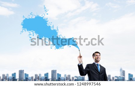 Horizontal shot of cheerful and young businessman in black suit gesturing and smiling while standing with blue coloured musical splash against cityscape view on background.