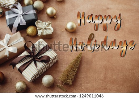 Happy Holidays text, handwritten golden sign at christmas stylish gift boxes with glitter tree and ornaments on rustic wooden background. Seasons greetings card