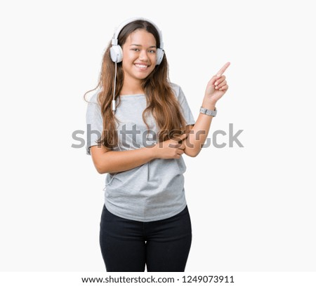 Young beautiful woman wearing headphones listening to music over isolated background with a big smile on face, pointing with hand and finger to the side looking at the camera.